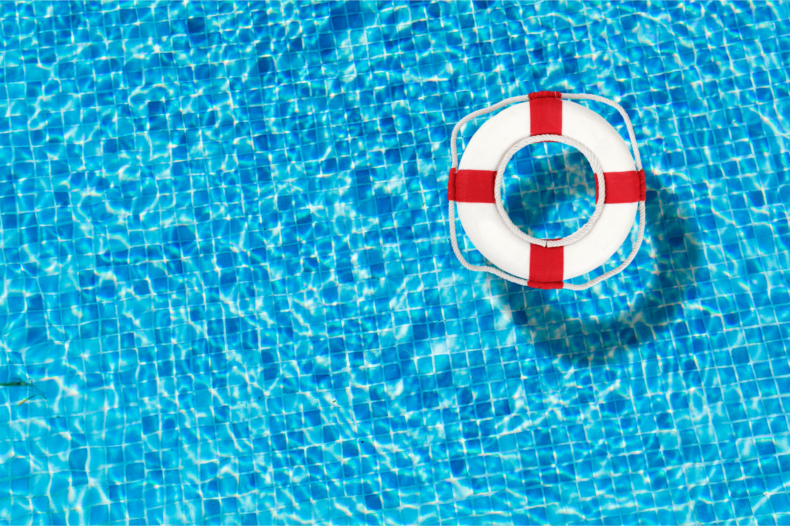 White lifebuoy with red bands floating on the surface of a blue tiled swimming pool.