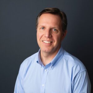 Smiling man in a light blue button-down shirt against a dark gray background (portrait)